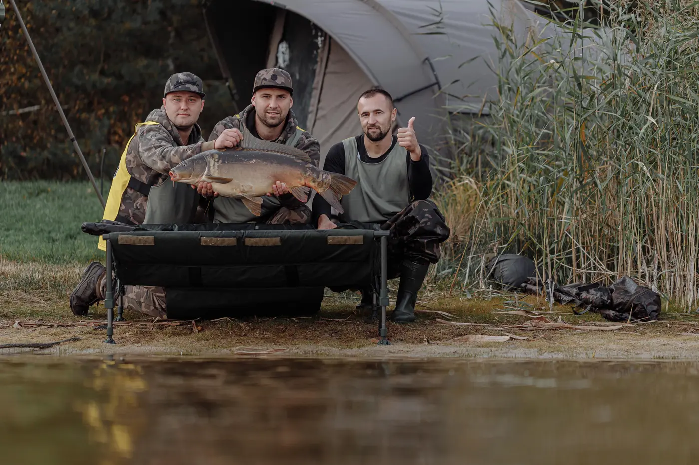 Three anglers with a carp on an unhooking mat next to a bivvy